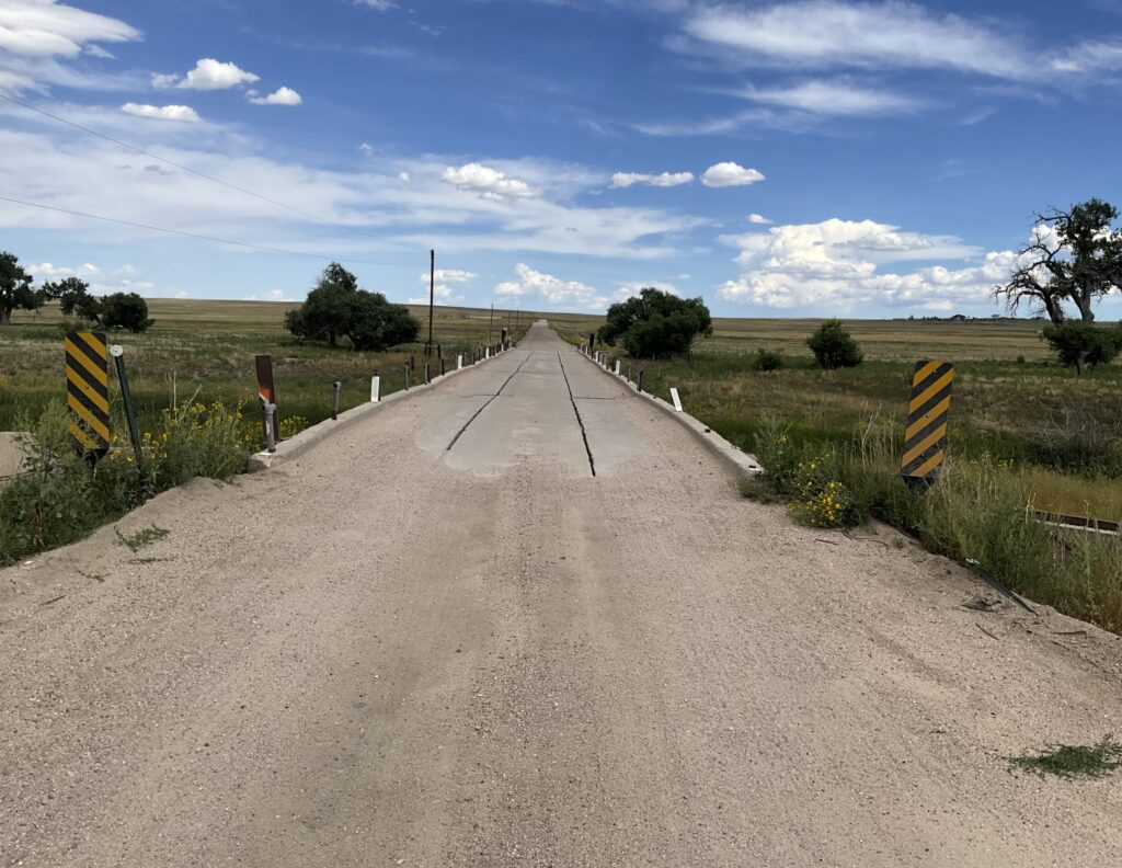 Gravel county road leading onto a narrow concrete bridge across open Elbert County prairie