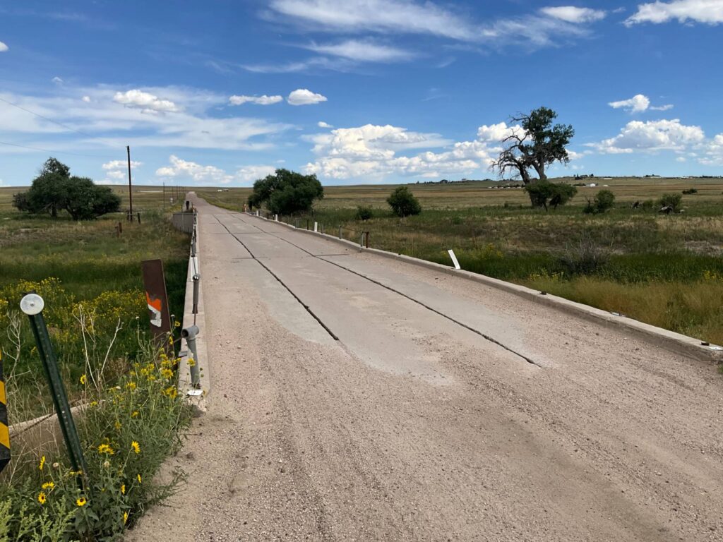 Narrow concrete bridge crossing open prairie with a cottonwood tree in Elbert County
