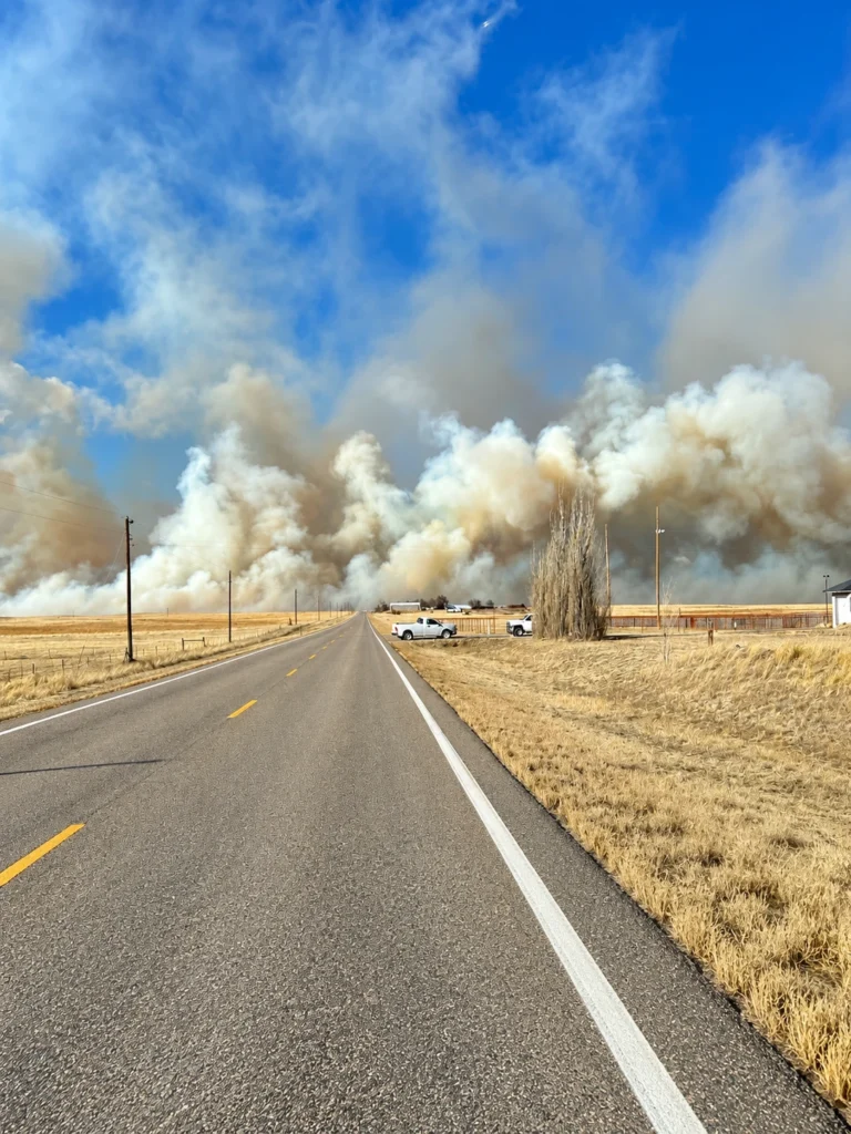 Wildfire smoke rising over dry grassland beside a rural two-lane road in Elbert County